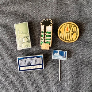 May include: Five enamel pins with different designs. The pins are all silver with gold accents. The pins feature images of a building, a palm tree, a circle with text, a building with text, and a building with text. The text on the pins reads "LENINGRAD" and "BELGOROD".