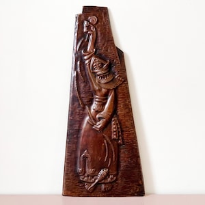 May include: A carved wooden wall art piece depicting a figure in traditional attire. The artwork is a rich brown color, with intricate details in the carving. The figure is holding an object aloft, with a detailed headdress and clothing.