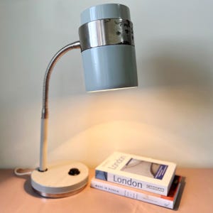 Puede incluir: Una lámpara de escritorio vintage con una pantalla cilíndrica azul claro y un cuello flexible plateado. La lámpara tiene una base blanca y un interruptor de encendido/apagado negro. Dos libros están apilados debajo de la lámpara.