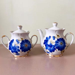 May include: A white ceramic teapot and sugar bowl set. Both pieces feature a blue floral design with gold trim. The teapot has a spout and handle, while the sugar bowl has two handles and a lid. The set is designed for serving tea.