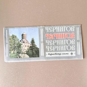May include: A rectangular travel brochure featuring a photograph of a church with a domed roof and trees. The brochure has text in Cyrillic script, with the word "Chernihiv" repeated multiple times. The brochure is on a light brown surface.