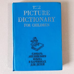 Op de afbeelding: Een blauw hardcover boek met de titel "THE PICTURE DICTIONARY FOR CHILDREN" in zilveren letters. Onder de titel staat een tekening van een kind en een boek. De onderkant van het boek bevat tekst in het Russisch.