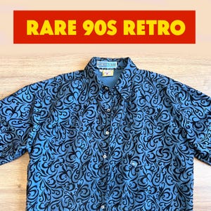 Può includere: Una camicia blu con un motivo a vortice nero. La camicia ha un colletto e una piccola tasca. Il testo "RARE 90S RETRO" è visualizzato in giallo su sfondo rosso sopra la camicia.