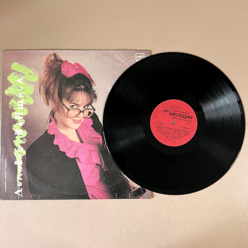 Puede incluir: Un disco de vinilo vintage y su portada. La portada presenta a una mujer con gafas, un lazo rosa y una camisa rosa con volantes. El disco es negro con una etiqueta roja con texto cir&iacute;lico.