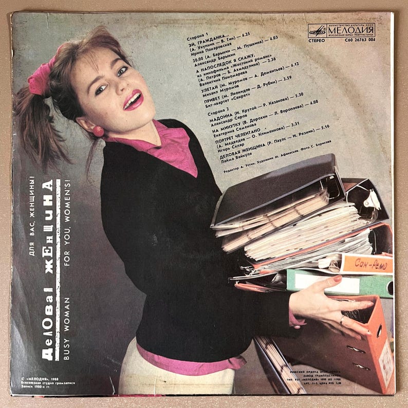 Puede incluir: Portada de &aacute;lbum de vinilo vintage con una mujer sosteniendo archivos, con texto en ruso e ingl&eacute;s: "Busy Woman" y "For You, Women's!" La portada del &aacute;lbum tiene una est&eacute;tica retro con una mujer con camisa rosa y chaqueta negra.