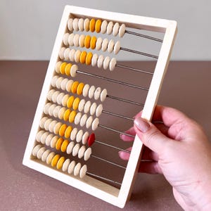 May include: A vintage abacus with a cream-coloured frame and rows of colourful beads. The beads are a mix of white, yellow, and one red bead. The abacus is held by a person.