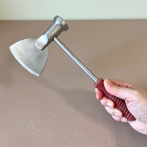 Puede incluir: Una herramienta de metal con cabeza de martillo y una cuchilla. La cuchilla es triangular y el mango es rojo. La herramienta se sostiene en una mano.