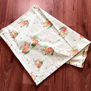 May include: A light green baby blanket with a repeating pattern of cartoon bees. The bees are orange and brown with green wings, and are resting on green leaves. The blanket is folded on a wooden surface.