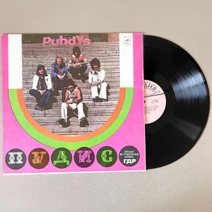 May include: A vintage vinyl record album cover featuring the band Puhdys. The cover has a pink background with green and orange concentric circles. The band members are seated on wooden steps. A black vinyl record is next to the album cover.