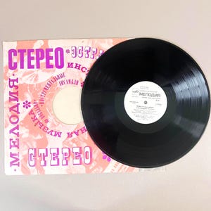 Puede incluir: Un disco de vinilo negro con una etiqueta blanca, colocado sobre su funda rosa y blanca. La funda presenta texto cirílico y la palabra "STEREO". Un artículo de música vintage.