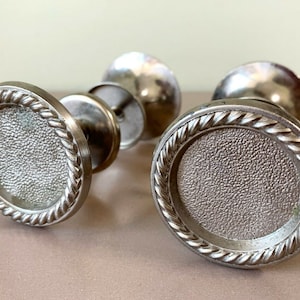 Puede incluir: Dos pomos de puerta plateados con un diseño de cuerda alrededor del borde. Los pomos tienen una superficie texturizada.