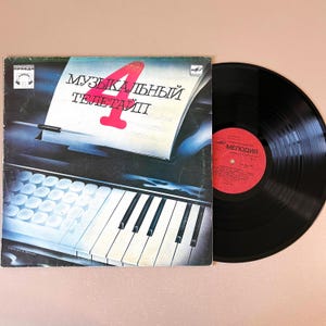May include: A vintage vinyl record with its cover. The cover features a piano keyboard illustration with the Russian text "4 Музыкальный Телетайп" in white and red. The record itself is black with a red label.