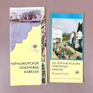 May include: Two vintage travel brochures. One brochure has a yellow background with the text "ЧЕРНОМОРСКОЕ ПОБЕРЕЖЬЕ КАВКАЗА" and a map illustration. The other brochure has a blue background with the text "ПО ЧЕРНОМОРСКОМУ ПОБЕРЕЖЬЮ КАВКАЗА (Сухуми-Сочи)" and a photo.