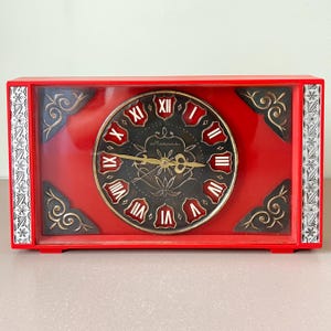 May include: A vintage red rectangular clock with a black clock face featuring gold hands and Roman numerals. The clock face is surrounded by a red frame with decorative gold accents and a clear plastic cover.