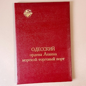 May include: A red, textured booklet with gold lettering and a gold emblem at the top. The text reads "ОДЕССКИЙ ордена Ленина морской торговый порт". The booklet appears to be a certificate or document.