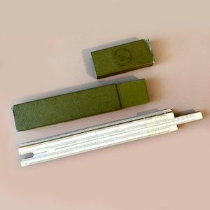 Op de afbeelding: Een vintage rekenliniaal met een witte behuizing en zwarte markeringen, vergezeld van een groene koffer en een kleinere groene doos. Een wiskundig rekeninstrument.