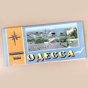 May include: Vintage postcard featuring a harbor scene with boats and buildings. The postcard has a blue and orange color scheme with the word "ОДЕССА" in gold lettering. The text "туристическая схема" is also visible.