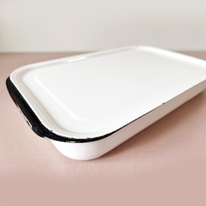 Puede incluir: Una bandeja rectangular blanca de esmalte con un borde negro. La bandeja tiene esquinas redondeadas y un borde ligeramente elevado. La bandeja está vacía y se encuentra sobre una superficie rosa. Probablemente se utiliza para servir o almacenar.