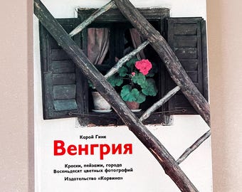 May include: A book cover featuring the title "Венгрия" in red, with text in Russian. The cover shows a window with dark shutters, a wooden frame, and a pot of pink flowers. The background is a light color.