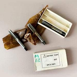 May include: A set of metric taps, with three pieces per set, presented with packaging. The taps are silver with ridged cutting edges. The packaging includes a box with the text "METRIC TAPS" and "3PCS/SET".