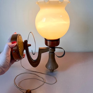 May include: Vintage wall sconce with a frosted glass globe and a decorative metal arm. The sconce has a pull chain with a wooden bead. The metal components have a bronze finish. The glass globe has a ruffled edge.