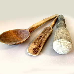 May include: Three antique wooden kitchen utensils: a large spoon, a spatula, and a pestle. The spoon has a deep bowl and long handle. The spatula is rectangular with a narrow handle. The pestle has a bulbous head and a cylindrical handle.