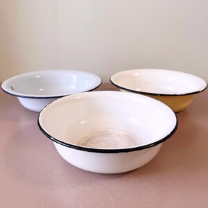 Könnte beinhalten: Drei Emaille-Schalen in Weiß- und Cremetönen, jede mit schwarzem Rand. Die Schalen sind auf einer rosa Oberfläche angeordnet und zeigen ihr schlichtes, klassisches Design. Diese Schalen sind ideal für Küche oder Heimdekoration.