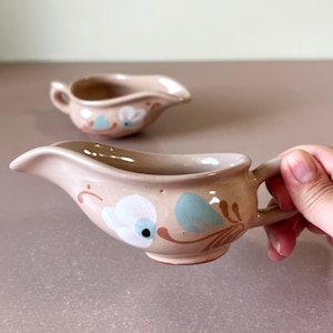 May include: Two light beige ceramic gravy boats featuring a hand-painted floral design in white, light blue, and brown. Each piece has a curved pouring spout and a small handle on the side, with a smooth, glossy finish on the exterior surface.