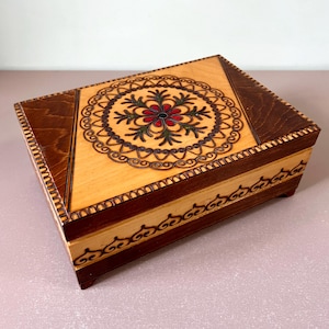 May include: A wooden box with a decorative floral design on the lid. The box is made of light and dark wood and has a hinged lid.