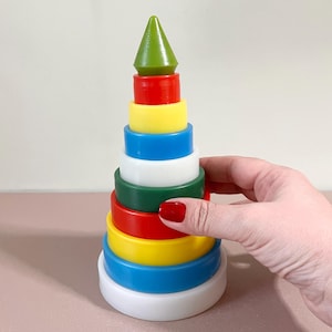 Puede incluir: Un juguete de apilar colorido hecho de plástico. El juguete tiene forma de cono y tiene una parte superior verde, anillos rojos, amarillos, azules, blancos y verdes. El juguete es un clásico para bebés y niños en edad preescolar.