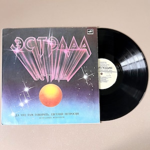 Puede incluir: Un disco de vinilo negro con una portada de álbum colorida que presenta el texto "ЭСТРАДА" y las palabras "ДА ЧТО ТАМ ГОВОРИТЬ. ЕВГЕНИЯ ПЕТРОСЯН ЭСТРАДНЫЕ МОНОЛОГИ". La portada del álbum también presenta una esfera naranja brillante y un cielo nocturno estrellado.
