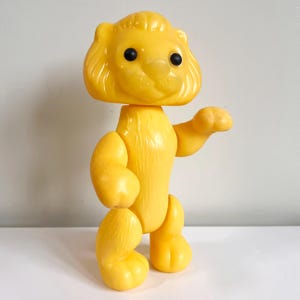 May include: A vintage yellow plastic lion toy with black eyes and articulated limbs. The lion has a mane and is posed with one arm raised. The toy is standing on a white surface against a neutral background.