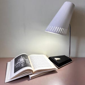 May include: A white conical desk lamp with a silver metal arm and a black trapezoidal base featuring a small white switch. The lamp is illuminated and positioned next to an open book with black and white printed text and a photograph of a building.