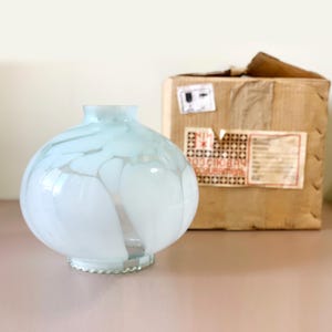 Può includere: Un paralume rotondo in vetro azzurro chiaro con venature bianche. Il paralume ha una base decorativa smerlata. Sullo sfondo c'è una scatola di cartone con etichette di spedizione.