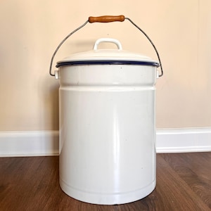 May include: A cylindrical white enamel canister with a matching lid and a dark blue rim. The container features a metal swing handle with a central wooden grip. The design is simple and functional, with a smooth finish and a sturdy construction.