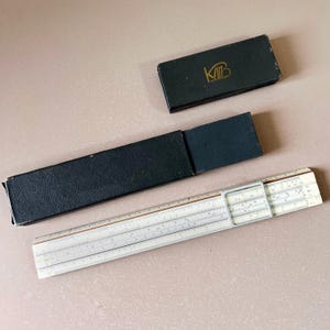 May include: A vintage slide rule with a black case and a smaller black box with gold lettering. The slide rule is off-white with black and brown markings. The smaller box has the letters "KMB" in gold.