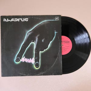 Puede incluir: Un álbum de vinilo vintage con una portada negra que presenta un gráfico de mano verde neón y azul. La portada del álbum tiene la palabra "ALEDPIUC" en una fuente futurista. El disco en sí es negro y parcialmente visible.