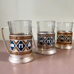 May include: A set of three ceramic mugs with a speckled, cream-colored glaze. Each mug features a different hand-painted floral design in shades of blue, green, and yellow. The mugs have a simple, rounded shape and a comfortable handle.