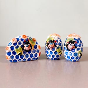 Może przedstawiać: Ceramiczny zestaw solniczki i pieprzniczki oraz serwetnik. Zestaw posiada wzór plastra miodu z niebieskimi i białymi sześciokątnymi wzorami. Każdy element ma uśmiechniętą twarz pszczoły i kolorowe kwiatowe akcenty.
