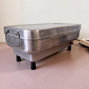 Peut inclure: Un appareil rectangulaire en aluminium brossé avec un couvercle à charnière et une poignée. L'appareil a quatre pieds noirs et un cordon d'alimentation. L'article est probablement un chauffe-plat ou un plat de service.