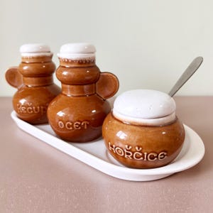 May include: A ceramic condiment set featuring three brown jars with white lids and handles, and a small spoon. The jars are labeled "Kecup", "Ocet", and "Horcice" and sit on a white, oval-shaped tray.