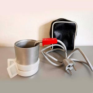 May include: A vintage electric travel cup with a stainless steel interior, white plastic base, and a red connector. A black leatherette case with white trim sits behind the cup. A grey power cord with a two-pin plug is also visible.