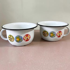 May include: Two white enamel mugs with black rims and handles. Each mug features colorful cartoon illustrations of a snail, frog, and butterfly. The mugs are likely vintage and add a touch of whimsy to any kitchen.