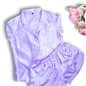 May include: A lavender satin pajama set with short sleeves and shorts. The top has a button-up front and a pocket on the left chest with a white embroidered letter "A" inside a wreath.