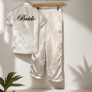 May include: Ivory satin pajama set with black trim, including a short-sleeved button-up shirt and matching pants. The shirt features the word "Bride" in elegant script on the back. The set hangs on a wooden hanger.