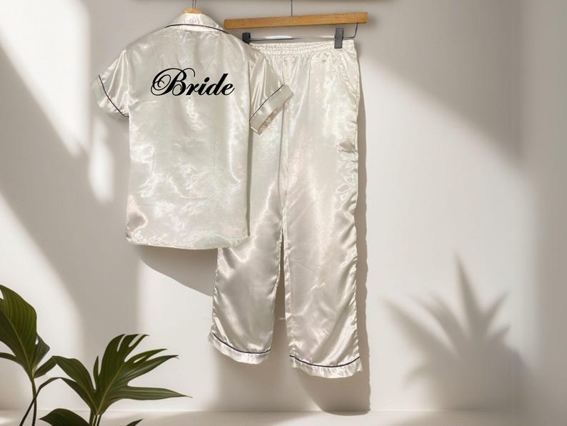 May include: Ivory satin pajama set with black trim, including a short-sleeved button-up shirt and matching pants. The shirt features the word "Bride" in elegant script on the back. The set hangs on a wooden hanger.
