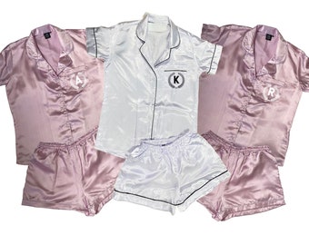 May include: Three satin pajama sets in pink, white, and pink. Each set includes a short-sleeved shirt and shorts. The shirts have a black trim and a small embroidered wreath with a letter in the center: A, K, and R.