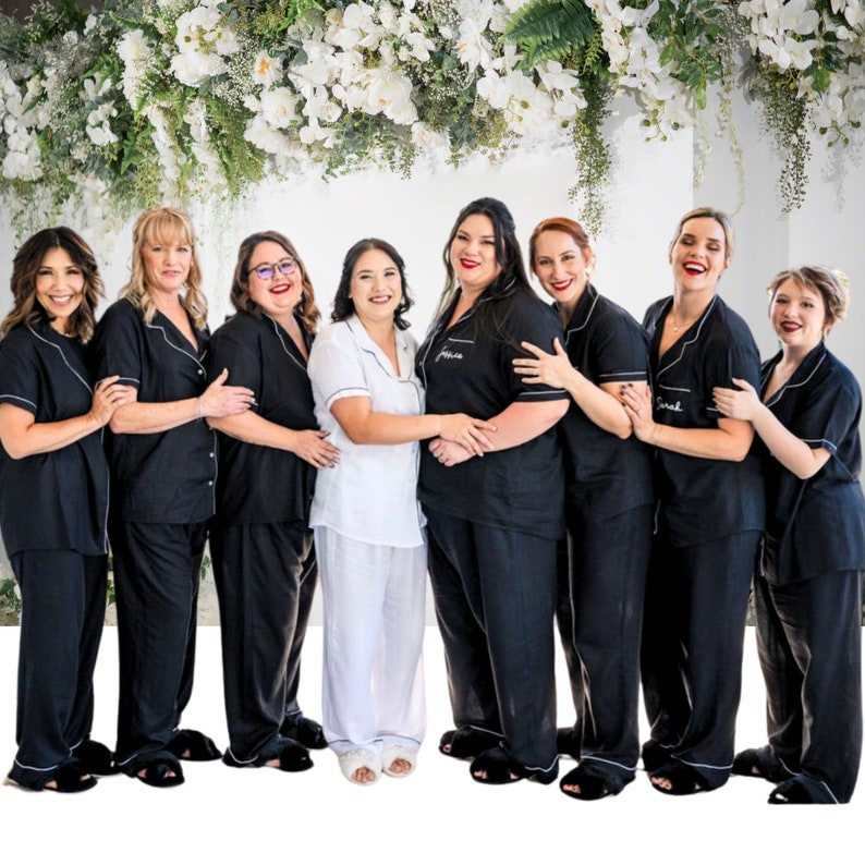 Pode incluir: Oito mulheres vestindo pijamas pretos com detalhes brancos e bordados personalizados. As mulheres est&atilde;o em fila, sorrindo para a c&acirc;mera. O fundo &eacute; uma parede branca com uma guirlanda de flores pendurada acima.
