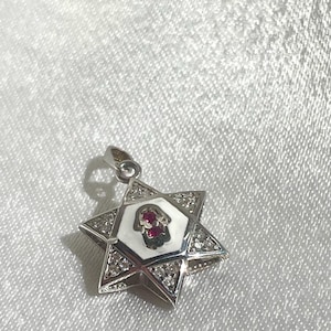 May include: A silver pendant in the shape of a Star of David with a white enamel center and a red gemstone. The pendant is decorated with small clear gemstones around the edges.