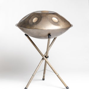Puede incluir: Un handpan de acero con acabado bronce y notas doradas se encuentra sobre un tr&iacute;pode de madera con una cadena que conecta las patas.